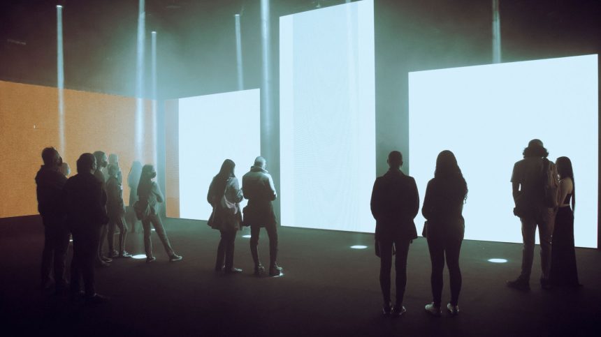 Visitantes exploram uma exposição de arte digital imersiva diante de grandes telas iluminadas.