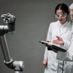 Pesquisadores de jaleco e óculos de proteção trabalham com um braço robótico em um laboratório.