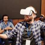Homem usa um headset de realidade virtual em ambiente interno, enquanto outras pessoas aparecem ao fundo.