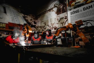 Três trabalhadores usando equipamentos de proteção atuam com robôs em uma oficina industrial de alta tecnologia.