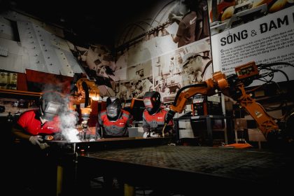 Três trabalhadores usando equipamentos de proteção atuam com robôs em uma oficina industrial de alta tecnologia.