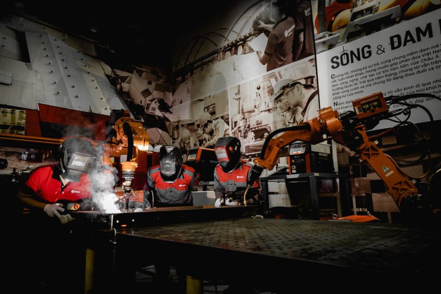 Três trabalhadores usando equipamentos de proteção atuam com robôs em uma oficina industrial de alta tecnologia.